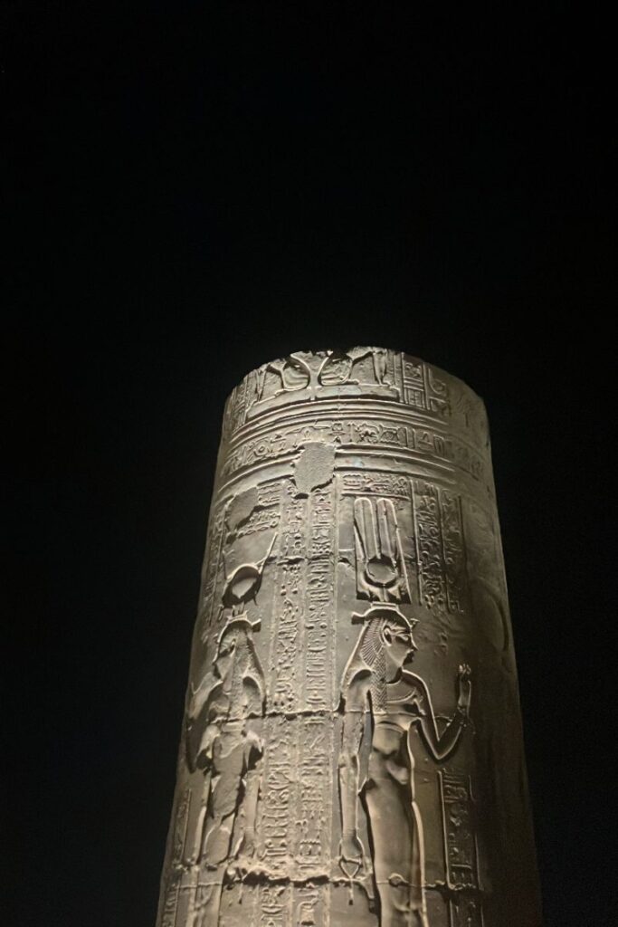 Illuminated stone column at night with hieroglyphs and carvings of Egyptian deities, part of Kom Ombo temple, dedicated to Sobek, the crocodile god of Egypt and protector of the Nile.