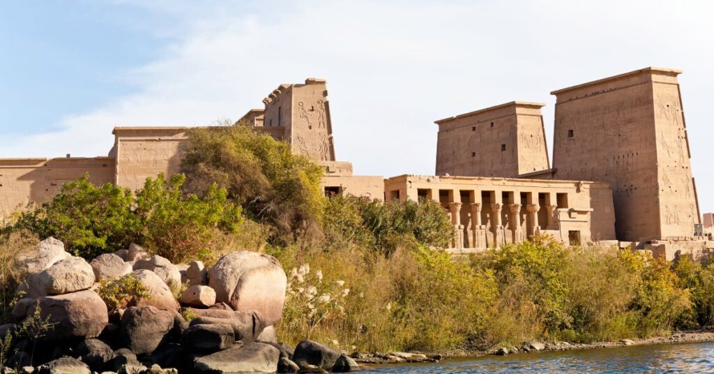 Scenic view of the Temple of Isis at Philae, Egypt, from across the Nile River on Philae Island, with lush vegetation in the foreground and the grand pylon towers and colonnaded halls of the ancient Philae temple complex rising behind them—an iconic landmark near Aswan Island that reflects deep layers of Philae temple history and meaning.