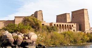 Scenic view of the Temple of Isis at Philae, Egypt, from across the Nile River on Philae Island, with lush vegetation in the foreground and the grand pylon towers and colonnaded halls of the ancient Philae temple complex rising behind them—an iconic landmark near Aswan Island that reflects deep layers of Philae temple history and meaning.