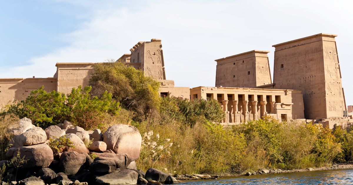 Scenic view of the Temple of Isis at Philae, Egypt, from across the Nile River on Philae Island, with lush vegetation in the foreground and the grand pylon towers and colonnaded halls of the ancient Philae temple complex rising behind them—an iconic landmark near Aswan Island that reflects deep layers of Philae temple history and meaning.