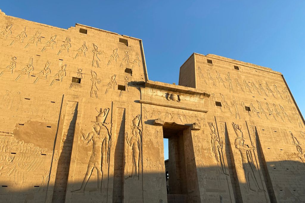 View of the grand pylon entrance gate of the Temple of Horus at Edfu, adorned with large bas-relief carvings of deities and pharaohs—a classic example of pylon architecture in Egypt.