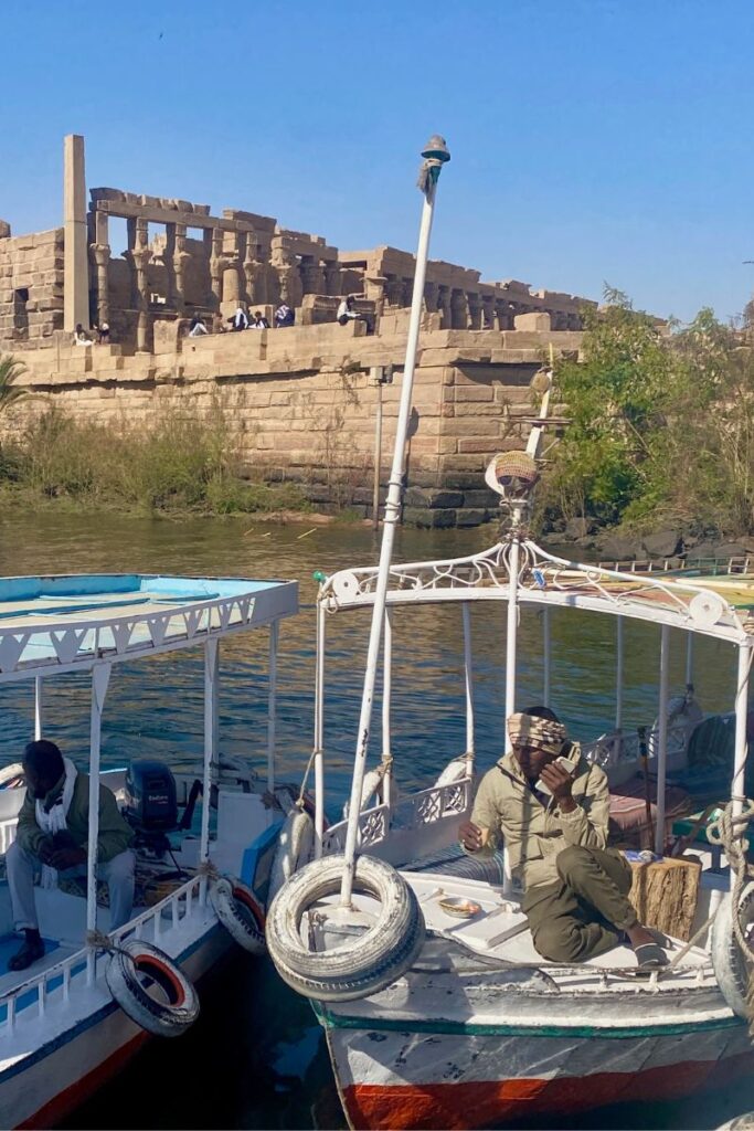 Tourists arriving at the Temple of Isis Philae by boat, with traditional feluccas docked below the grand sandstone ruins on Philae Island, Egypt.