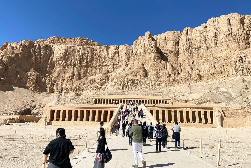 Tourists walk up the grand entrance of the Mortuary Temple of Hatshepsut in Luxor, surrounded by dramatic desert cliffs. A real-time snapshot for “is Egypt safe” and why to visit Egypt for its archeological wonders.