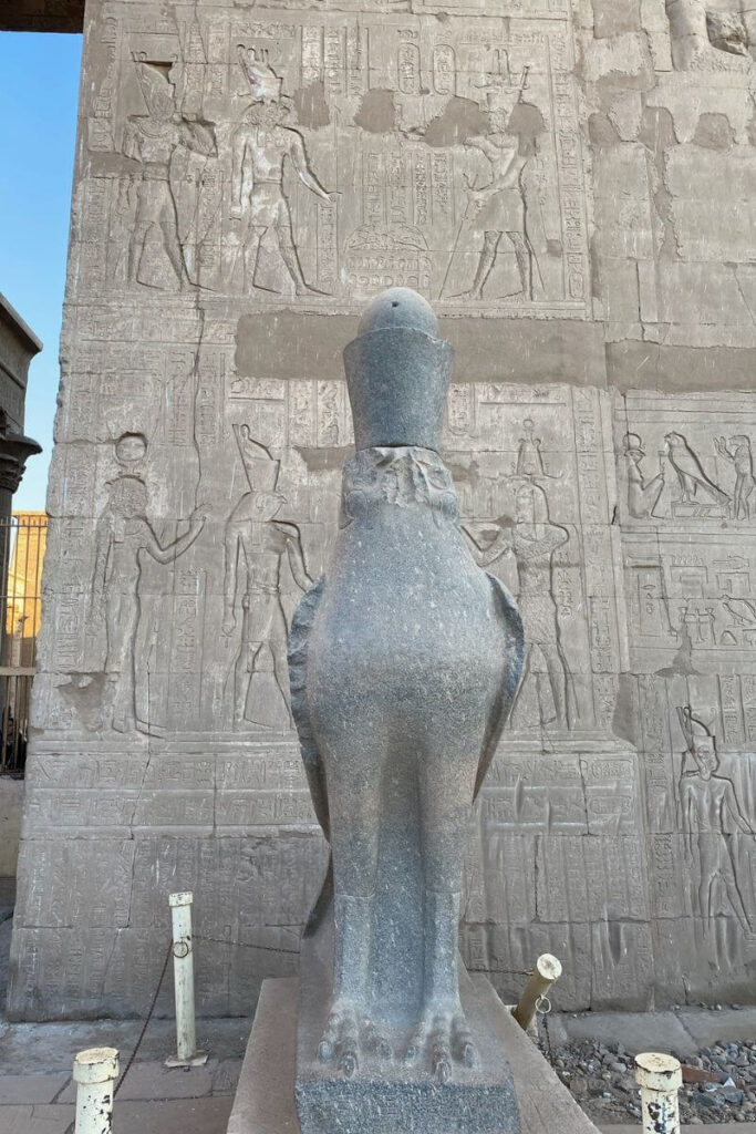 Statue of the falcon-headed god Horus wearing the double crown, standing outside a richly inscribed temple wall—perfect for visualizing Horus facts and his symbolic power in Egyptian temple art.