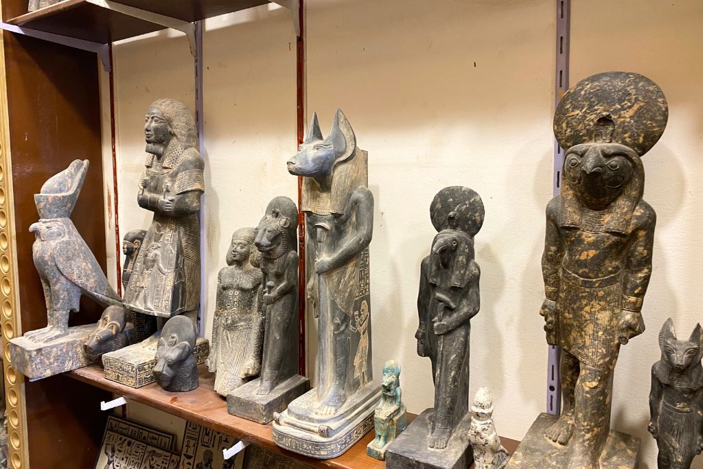 A collection of stone statues representing Egyptian gods and pharaohs, including Anubis, Horus, and Sekhmet, displayed on wooden shelves. A compelling look at why visit Egypt for its deep mythological history and culture.