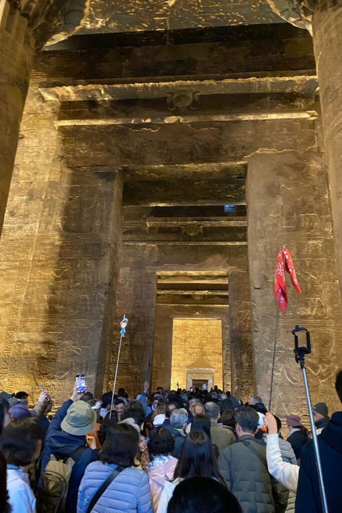 Large crowd of tourists moves through the dimly lit, hieroglyph-covered columns inside the Egyptian temple at Edfu—highlighting its popularity and rich atmosphere for those wondering why is the Temple of Horus at Edfu important.