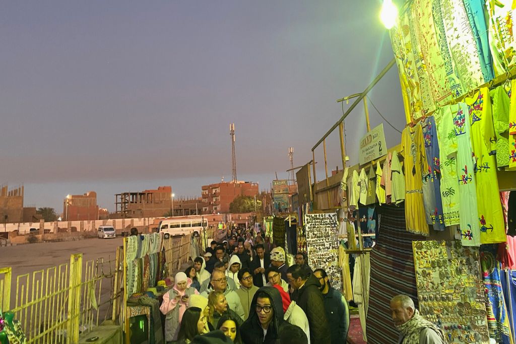 A long line of visitors passes vibrant market stalls selling souvenirs and clothes, a bustling stop en route to the famous temples in Egypt like the Edfu Temple.