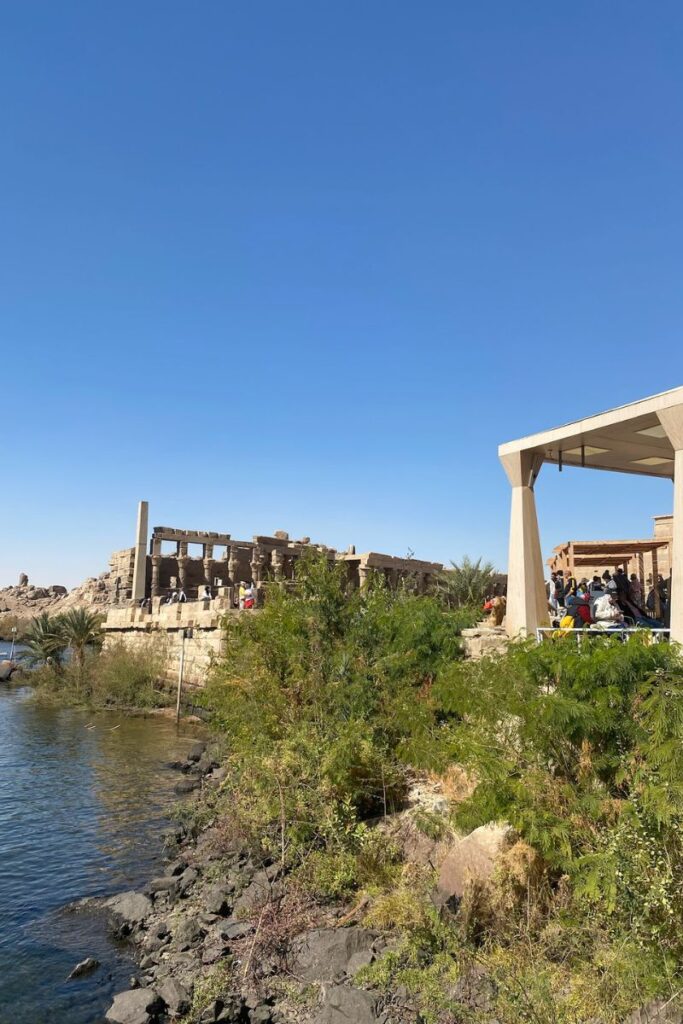 Tourists gather at the entrance of the Temple of Isis Philae, near the lush, riverside dock area of Philae Island Egypt, where boats arrive from Aswan to explore this historic site.