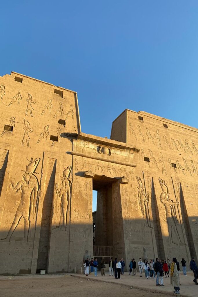Wide shot of a massive ancient Egyptian temple adorned with carvings and tourists at the entrance. Evokes tourism in Luxor and highlights interest in Uber in Luxor Egypt or comparing travel modes to Uber Egypt.