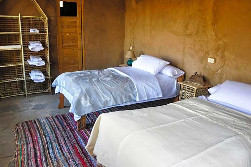 Interior of a rustic-style twin room at a boutique hotel in Aswan, featuring clay walls, a colorful rug, and open shelving with towels — a charming place to stay for travelers asking where to stay in Aswan Egypt.