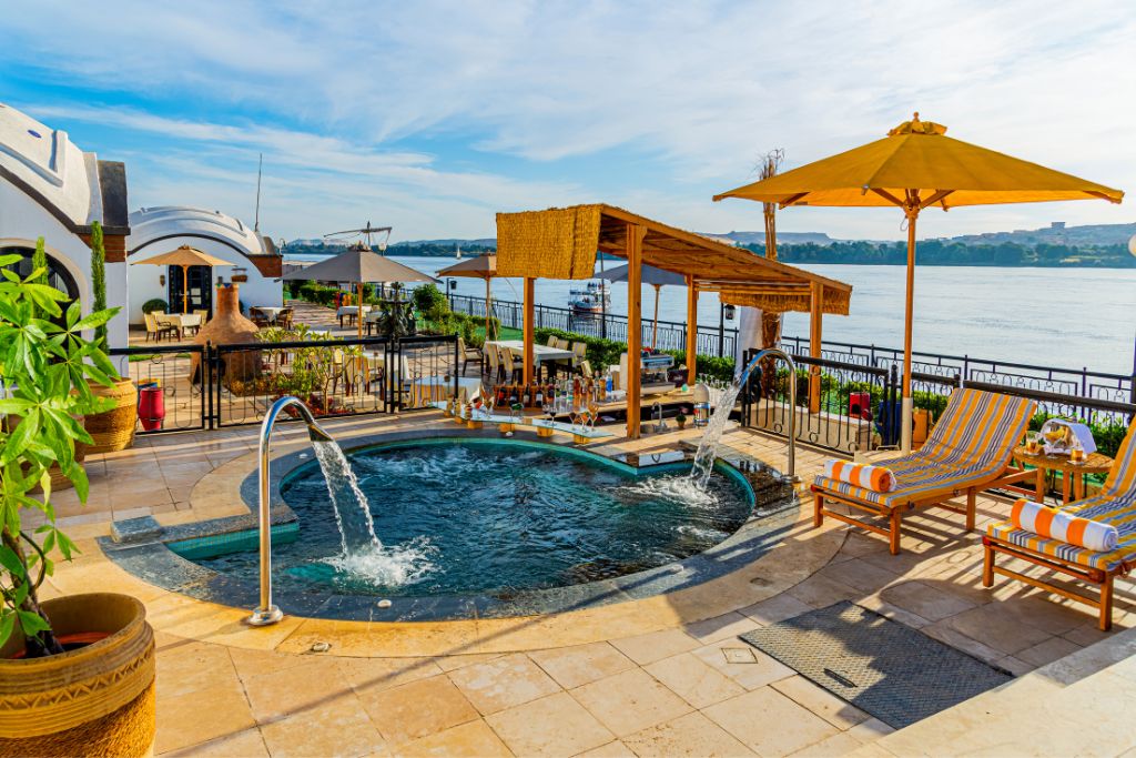 Riverside pool deck at a luxury hotel in Aswan, Egypt, featuring striped lounge chairs, an open-air bar, and a private jacuzzi with Nile River views — an ideal setting when deciding where to stay in Aswan Egypt and a prime example of 5-star Aswan hotels.