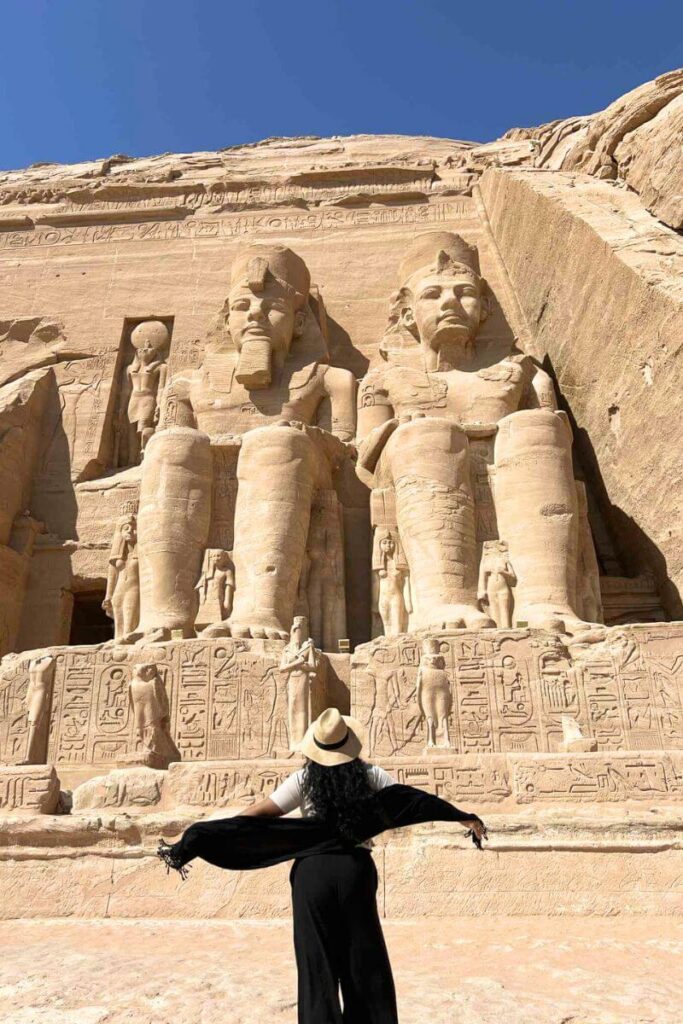 A woman in a wide-brimmed hat stands with arms outstretched in front of the colossal seated statues of Ramses II at Abu Simbel Temple, carved directly into the sandstone cliffs. The scene captures the grandeur of a day trip to Abu Simbel from Aswan under a clear blue sky.