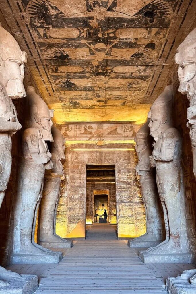 Interior view of Abu Simbel Temple in Egypt showing a corridor lined with towering statues and detailed wall carvings, illuminated with warm lighting. The long hallway leads to a shrine deep within the temple, a highlight on any Abu Simbel guide.