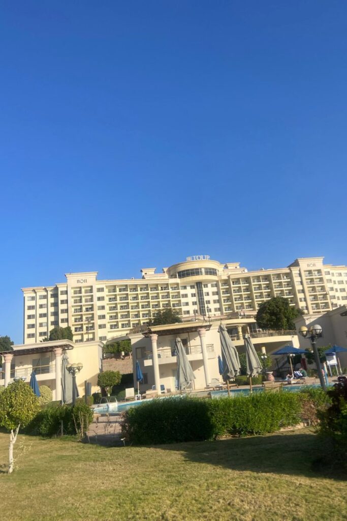 Wide-angle view of a luxury hotel in Aswan, Egypt, showing the main building with balconies, poolside umbrellas, and trimmed greenery under a bright blue sky — a popular choice for travelers searching for Aswan hotels or luxury hotels in Egypt.
