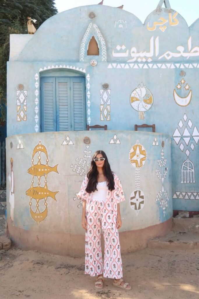A woman stands in front of a pale blue Nubian house adorned with white and gold tribal patterns and Arabic script in a Nubian village in Aswan. The traditional architecture and decorative murals highlight local culture and craftsmanship.