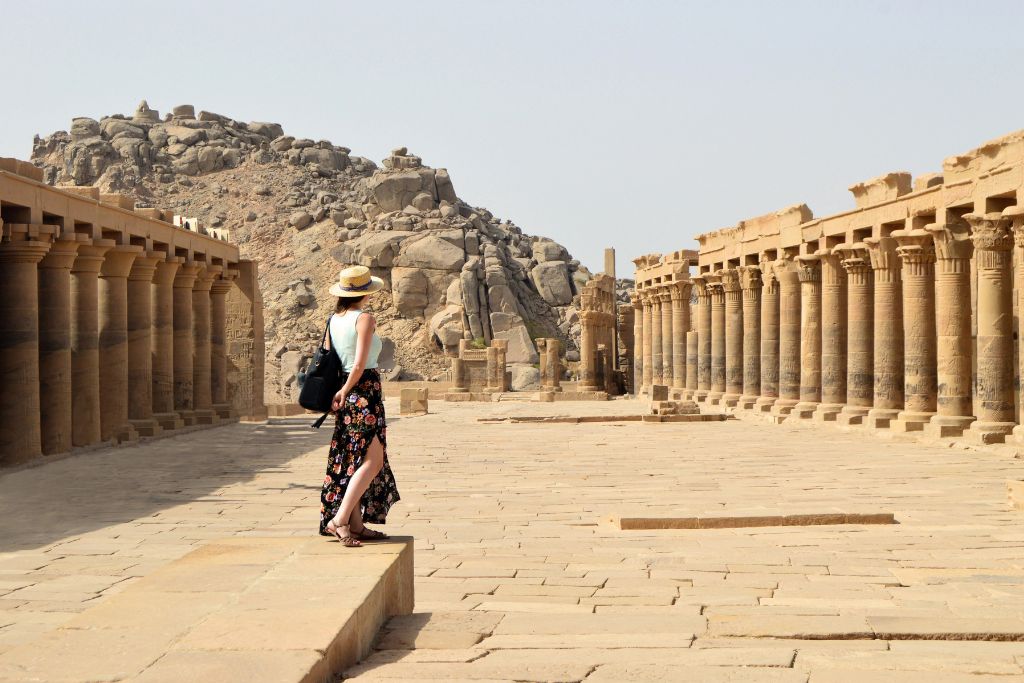 A woman walks between two long rows of sandstone columns at the Philae Temple near Aswan, surrounded by rocky desert terrain. This historic site is a must-see for travelers exploring ancient landmarks in southern Egypt.