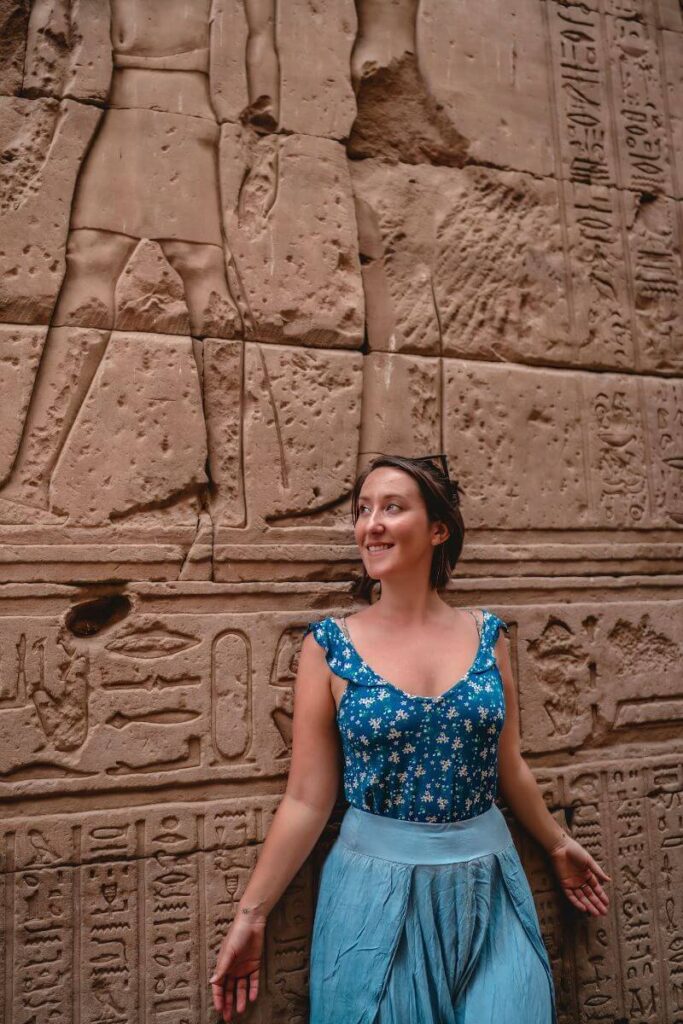 A woman in a blue floral top and light blue skirt poses against a sandstone wall carved with detailed hieroglyphs and partial pharaonic figures inside Philae Temple Egypt. This ancient Egyptian temple is one of the top historical attractions in Aswan and is dedicated to the goddess Isis.