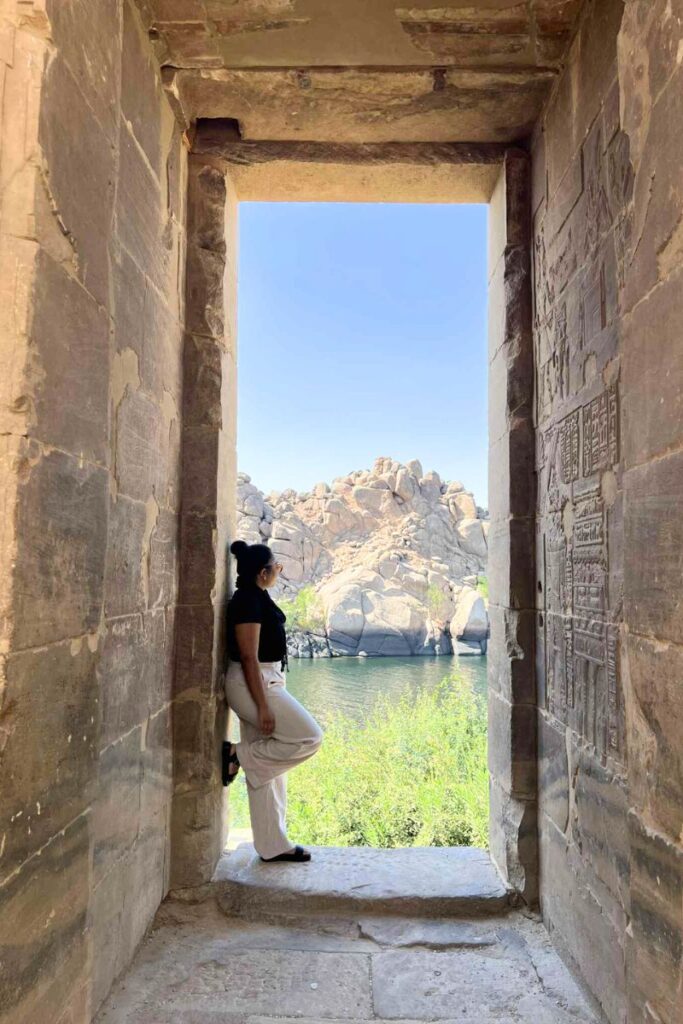 A woman in black and white stands in the doorway of Philae Temple in Aswan, looking out toward the Nile River and granite rock formations. The Temple of Isis offers serene views and is one of the most photographed ancient Egyptian temples in southern Egypt.