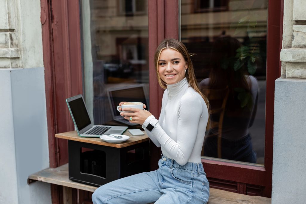 Woman sitting outside a café window holding a coffee beside her laptop, confidently eating alone at a restaurant. This relaxed solo restaurant scene shows how to enjoy solo dining and build confidence eating alone while working remotely.