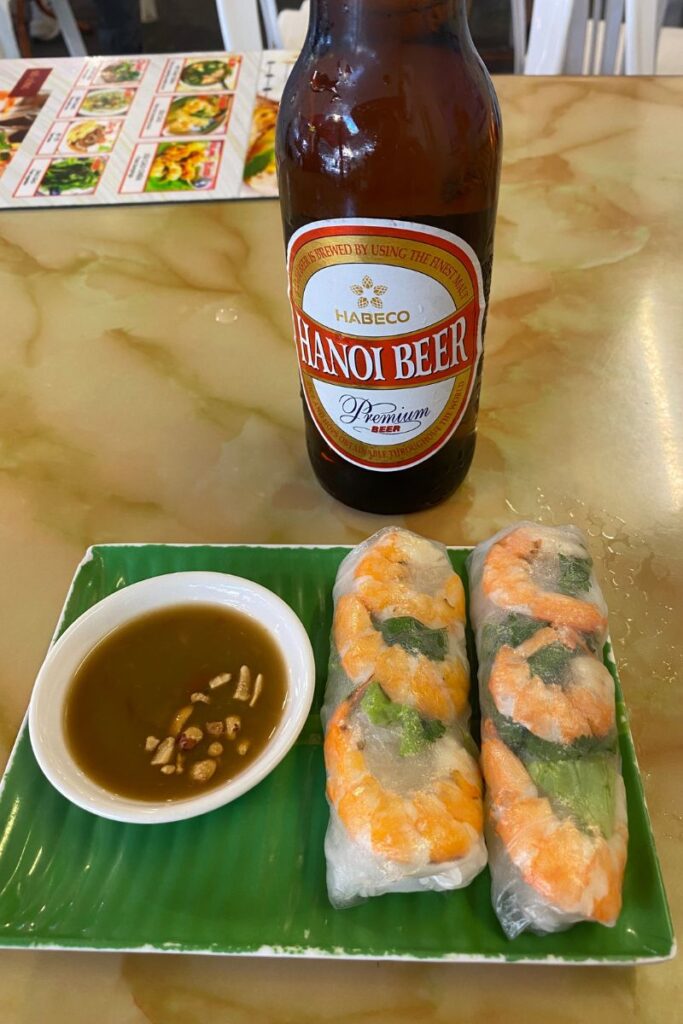 A woman in a café enjoys Vietnamese spring rolls with dipping sauce, accompanied by a cold Hanoi beer. A relaxed and relatable depiction of eating alone while traveling or enjoying a solo lunch.