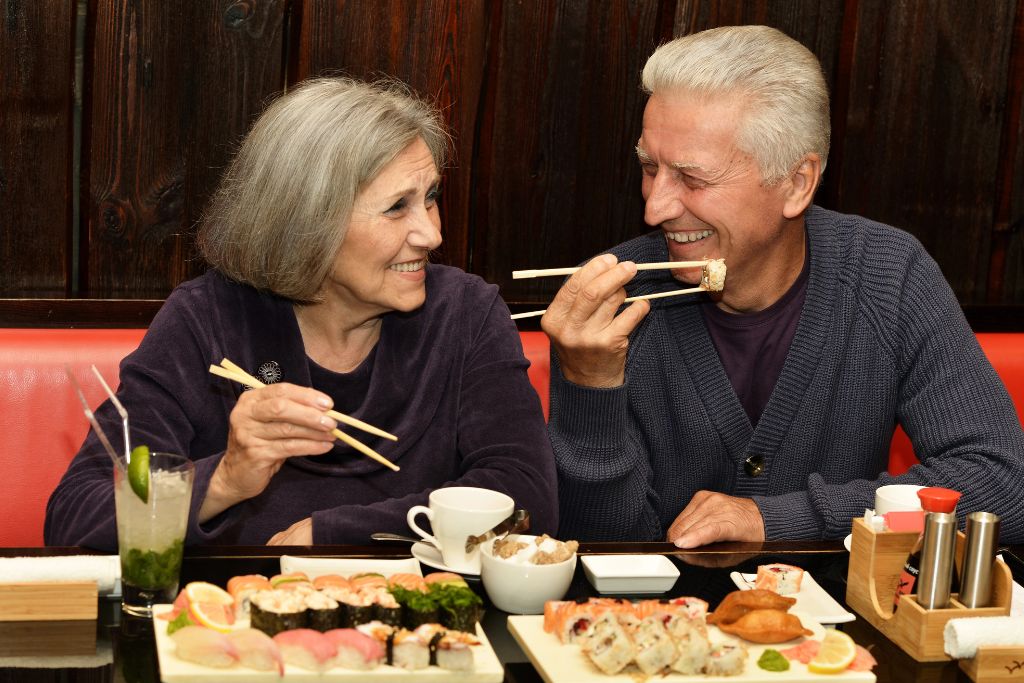 An older couple shares sushi joyfully, each using chopsticks, emphasizing companionship — a contrasting moment perfect for highlighting the courage it takes to eat alone in a restaurant.