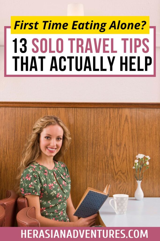 Smiling woman in a floral dress reads a book at a café table with coffee, next to the caption "First time eating alone in a restaurant: 13 Solo Travel Tips That Actually Help." A great visual for content on how to eat out alone, tips for eating alone as a woman, or first-time solo restaurant visits.
