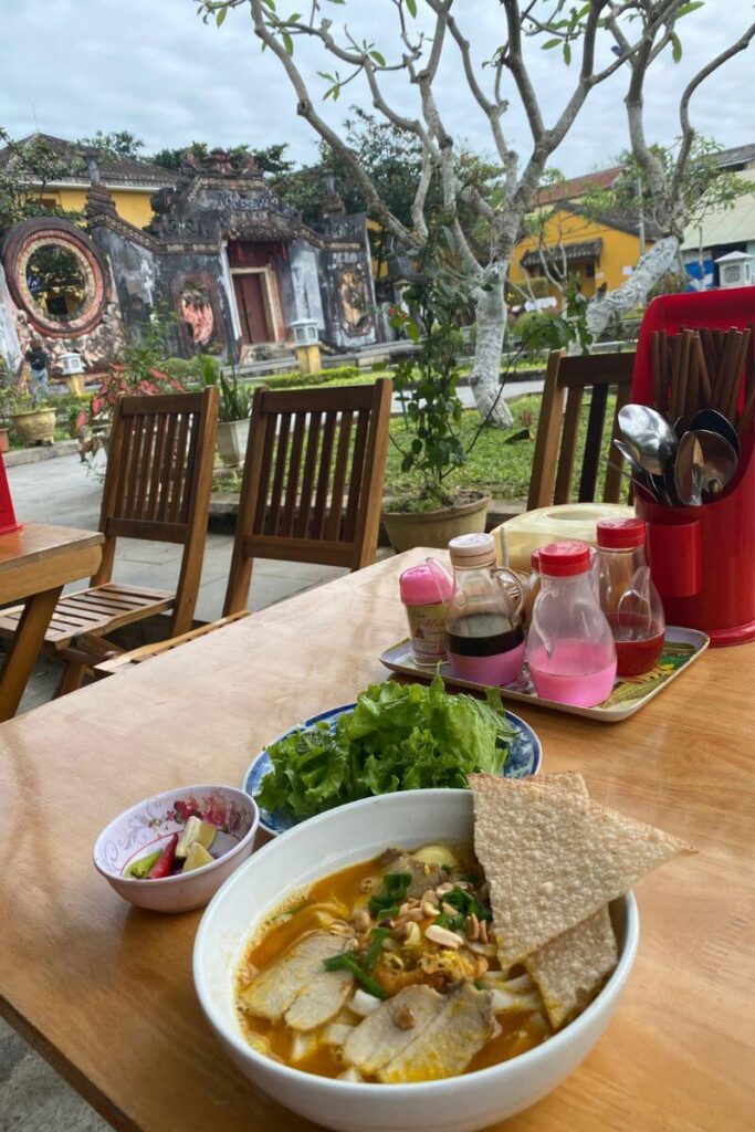 A serene outdoor setting in front of a historic building features a bowl of noodle soup, fresh greens, and condiments on a wooden table — ideal for solo travel eating alone or enjoying a meal solo while exploring.