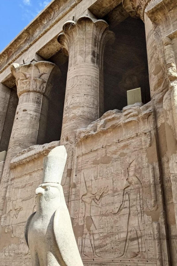 Close-up of the Temple of Horus at Edfu, Egypt, showing massive columns engraved with ancient Egyptian hieroglyphs and a stone statue of the falcon-headed god Horus in the foreground. This well-preserved structure is one of the top temples to visit in Egypt.