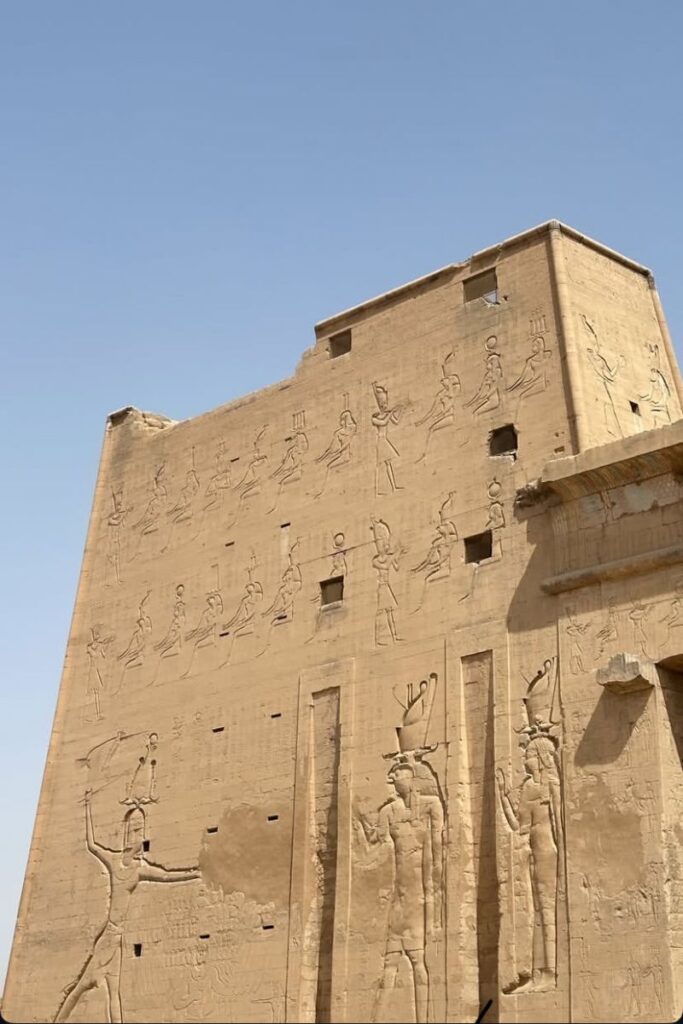 Side view of the towering sandstone walls of Edfu Temple in Egypt, carved with large-scale hieroglyphics and scenes of ancient rituals, including depictions of pharaohs and gods. The detailed reliefs showcase the artistry of the Temple of Horus, one of the best-preserved Egyptian temples.