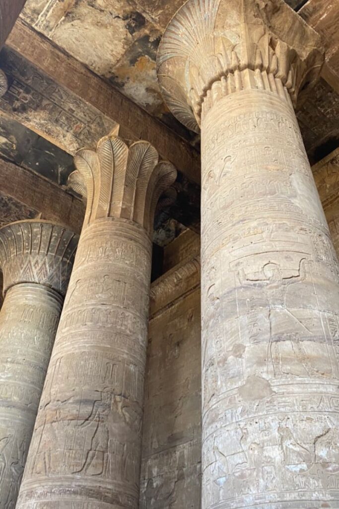 Interior shot of Edfu Temple in Egypt, highlighting towering sandstone columns with intricate hieroglyphic carvings and lotus-shaped capitals under a darkened stone ceiling. A must-see Egyptian temple known for its preserved Ptolemaic architecture.