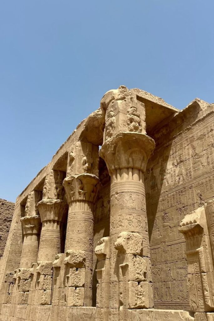 Exterior view of the Temple of Horus at Edfu under a bright blue sky, showing rows of detailed, carved sandstone columns and walls covered in ancient hieroglyphics. One of the most iconic and best-preserved temples in Egypt.
