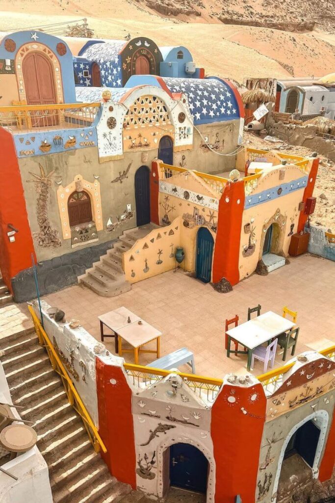 An aerial view of a vibrant courtyard surrounded by traditional Nubian houses with domed roofs and star-patterned murals in bold red, blue, and beige tones. This scene showcases authentic Nubian architecture in a village near Aswan, Egypt.