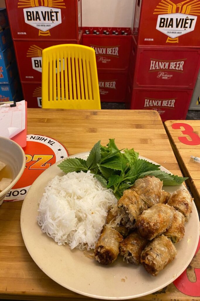 A vibrant Vietnamese dish of fried spring rolls, herbs, and vermicelli noodles on a wooden table, placed in front of stacked beer crates. This scene captures casual solo dining and the relaxed vibe of eating alone at a restaurant.