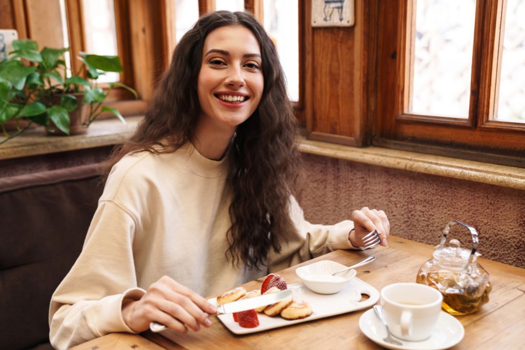 A woman smiles while dining solo at a cozy wooden restaurant table with a plate of biscuits, strawberries, and a teapot. This image conveys how to enjoy solo dining and the ease of eating alone at a restaurant.
