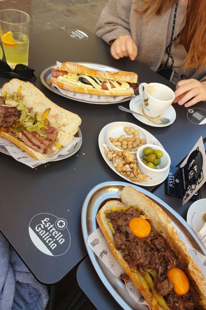 Outdoor café table with gourmet sandwiches topped with grilled meats and fried eggs, served with coffee and snacks. Captures a relaxed moment of eating in a restaurant alone or enjoying solo dining on a European terrace.