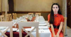 A woman in a red dress sits alone at a long restaurant table set for many guests, looking weird and awkward. Captures the feeling of dining alone as a woman and the question of whether going to a restaurant alone is unhealthy or empowering.