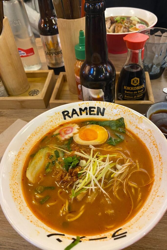 A vibrant bowl of ramen with egg, vegetables, and noodles sits surrounded by soy sauce, beer, and hot sauce bottles in a cozy restaurant. A perfect setting for solo eating or tips for dining alone in a bustling ramen bar.