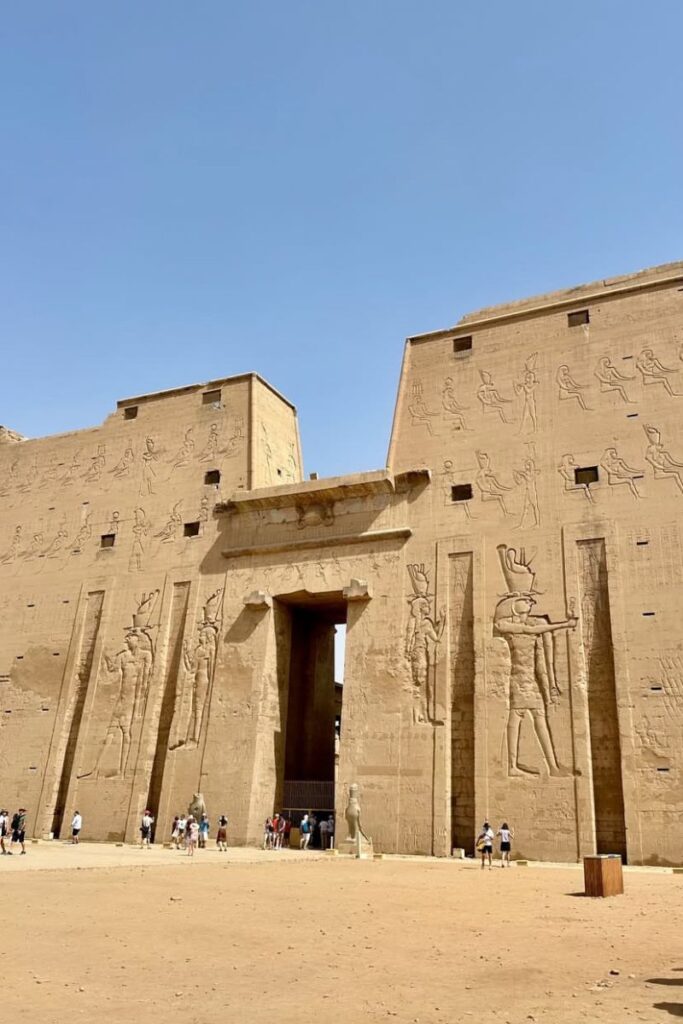 Wide-angle view of the grand entrance to the Edfu Temple in Egypt, featuring two massive pylons etched with hieroglyphics and figures of Horus and the Pharaoh. Tourists walk across the temple’s forecourt, emphasizing its scale and importance as a major site among temples to visit in Egypt.