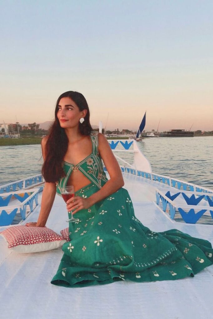 A woman in a green embroidered dress relaxes on a felucca boat with a glass of wine during sunset on the Nile River in Aswan. Felucca rides are one of the top things to do in Aswan for a peaceful view of the Egypt Nubian village scenery.