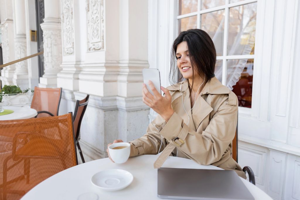 Woman smiling at her phone with coffee at a patio table, confidently going out to eat alone. A strong visual for dining alone as a woman and overcoming fear of going alone to a restaurant.