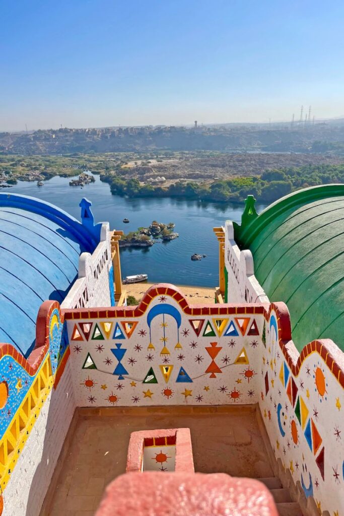 View from the rooftop of a brightly painted building in a Nubian village in Egypt, looking out over the Nile and Elephantine Island, emphasizing the blend of artistic heritage and natural beauty.