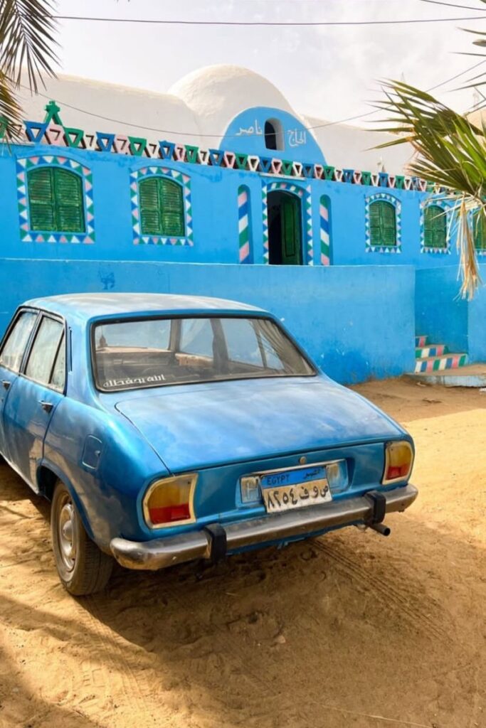 A vintage blue car parked outside a vividly painted blue Nubian home with green shutters and geometric border designs. This charming street scene is typical of daily life in a Nubian village in Egypt.