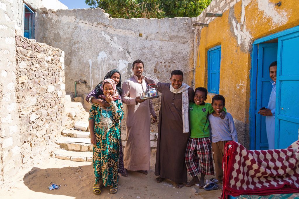 Smiling local family standing outside traditional stone and mud-brick houses with blue doors in a Nubian village in Aswan, Egypt, representing daily life and community in Nubian villages.