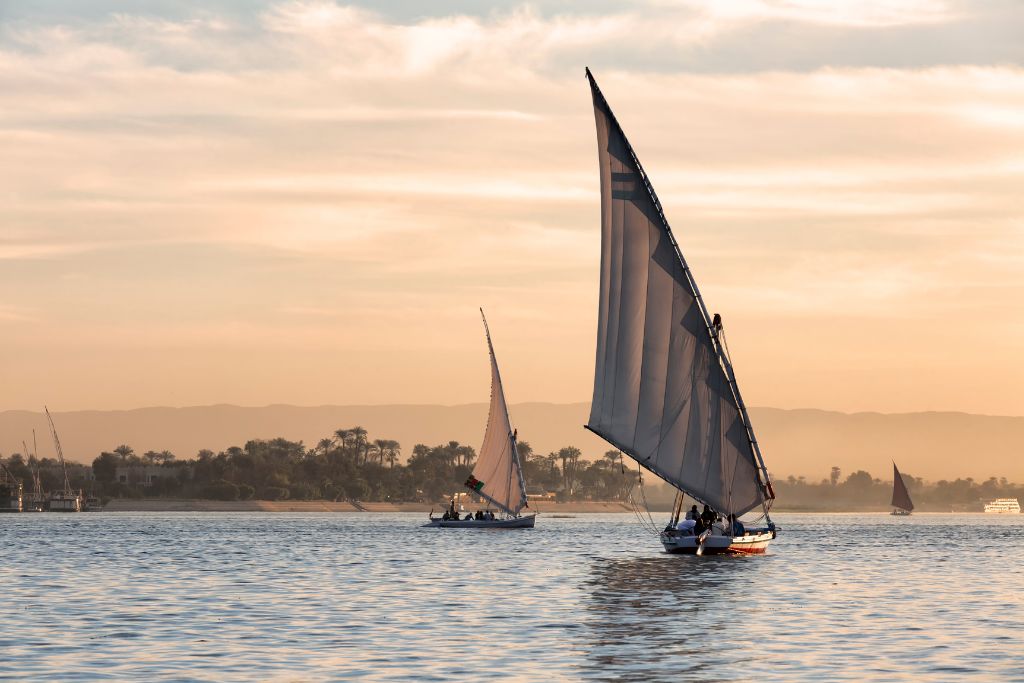 Traditional white-sailed feluccas glide along the Nile River at sunset, with a soft pastel sky and distant palms in the background. Felucca sailing is a relaxing and iconic experience when visiting the Nubian village Aswan region.