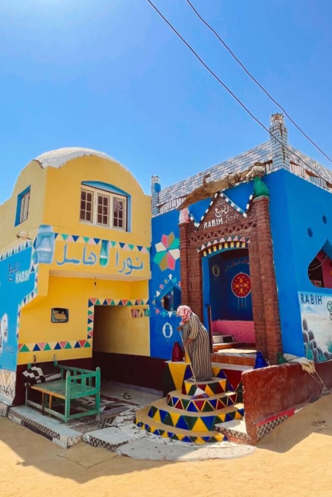 Two colorful Nubian houses painted in bright yellow, blue, and red with geometric patterns and Arabic writing, set under a clear blue sky. A woman in traditional dress sweeps the steps, offering a glimpse into everyday life in a Nubian village in Aswan.