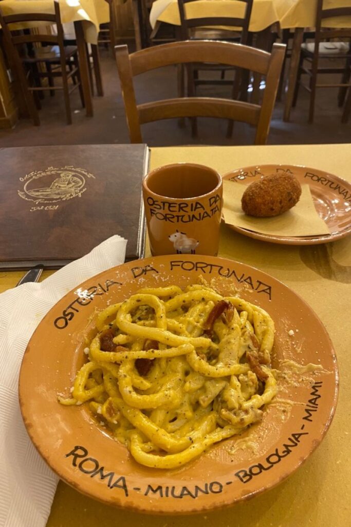 A plate of traditional Italian pasta carbonara with crispy guanciale, served beside a croquette and espresso cup at a rustic trattoria. Great for showcasing solo dinner ideas or dining alone while traveling through Italy.