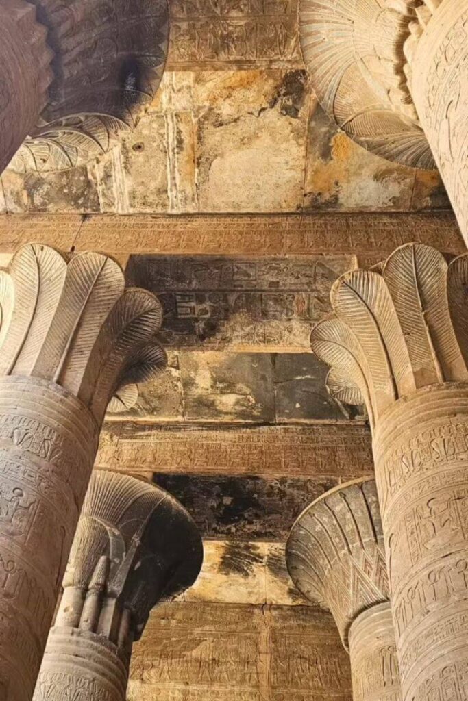 Upward view of the ceiling inside Edfu Temple in Egypt, framed by massive lotus-capped columns and detailed inscriptions. The preserved temple architecture offers a glimpse into ancient Egyptian religious art and design.