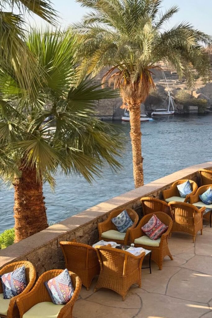 Rattan chairs with colorful cushions overlook the Nile River on a sunny terrace near Philae Temple in Aswan. With palm trees and scenic river views, this peaceful setting offers a relaxing spot close to one of the most famous archaeological sites in Egypt.