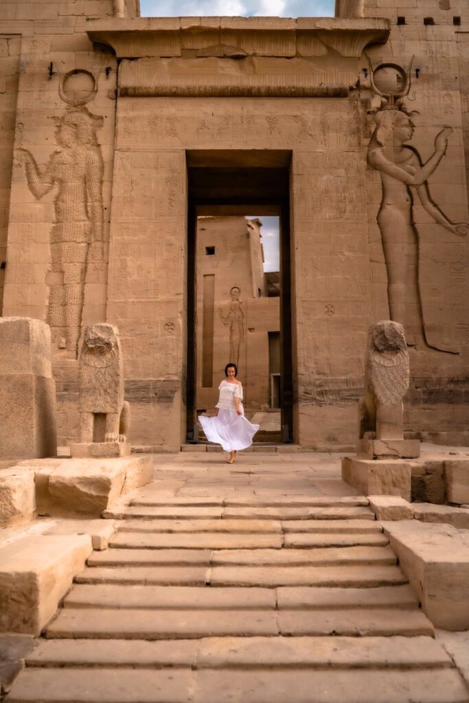 A woman in a flowing white dress walks up the grand staircase of the Temple of Isis at Philae, framed by colossal carvings of ancient Egyptian goddesses and twin lion statues. This temple complex is one of the most beautiful places to visit in Aswan, Egypt.