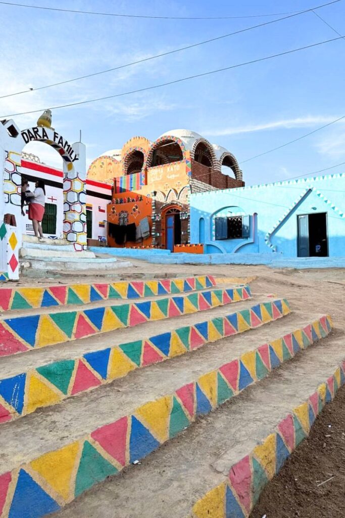 Brightly painted stairs lead to a colorfully decorated Nubian guesthouse with signs reading "Dara Family" and "Soma Kato," near the Philae Temple complex in Aswan. These vibrant villages reflect the local culture and are often visited by tourists exploring southern Egypt.